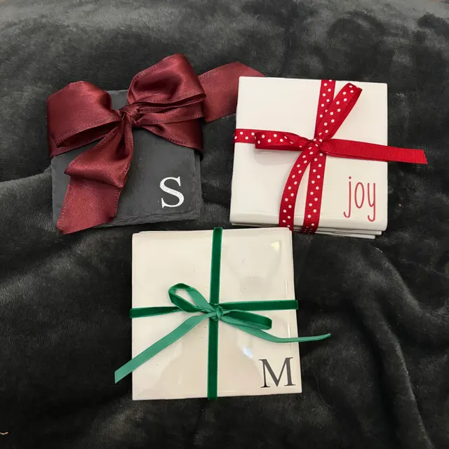 Three square coasters on a dark plush surface — a slate S, a ceramic joy and a ceramic M, each tied with a ribbon