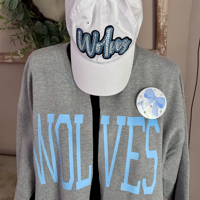 Wolves spirit set — white cap with a glittery blue Wolves script, a heather grey WOLVES sweatshirt, and a round EMS bow badge