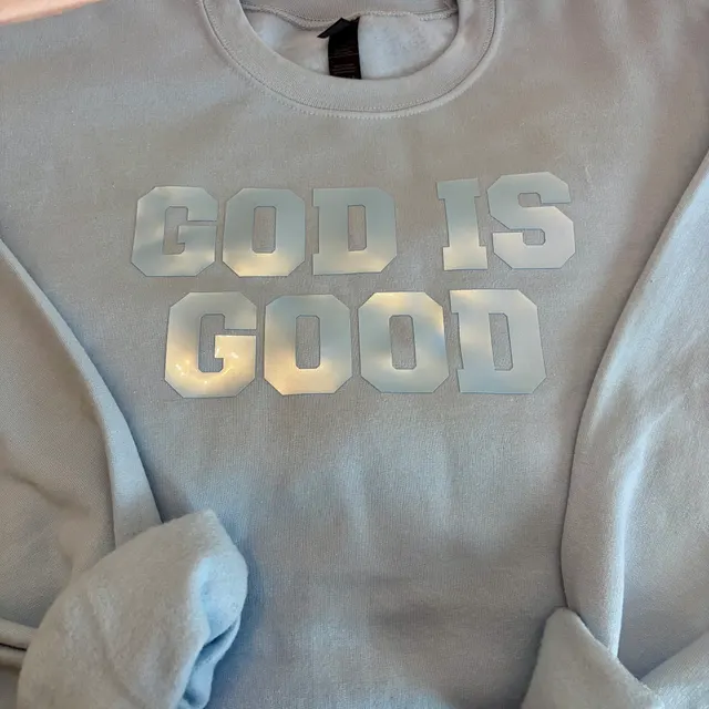 Light blue crewneck sweatshirt with GOD IS GOOD in blocky pale blue letters across the chest, sleeves folded