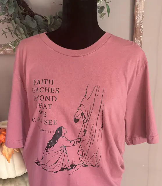 Dusty pink t-shirt on a black mannequin printed FAITH REACHES BEYOND WHAT WE CAN SEE with HEBREWS 11:1, beside a simple line drawing of two figures