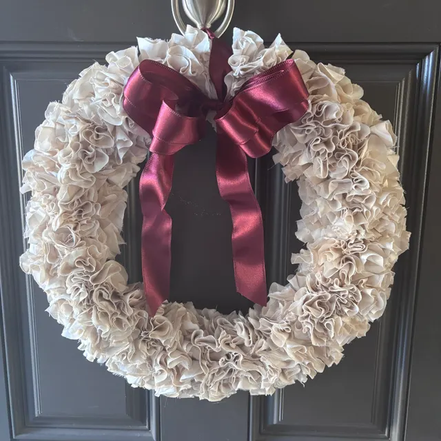 Cream fabric wreath with ruffled petals and a hand-tied burgundy satin bow, hanging on a dark grey panelled door